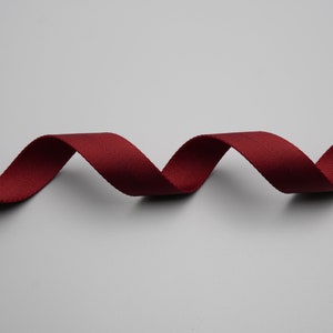 Peut inclure: Un ruban de couleur bordeaux, tiss&eacute; avec un motif diagonal subtil, ondule sur un fond gris. La texture du ruban est visible, sugg&eacute;rant un mat&eacute;riau durable. Le ruban est probablement utilis&eacute; pour l'artisanat, la couture ou &agrave; des fins d&eacute;coratives.
