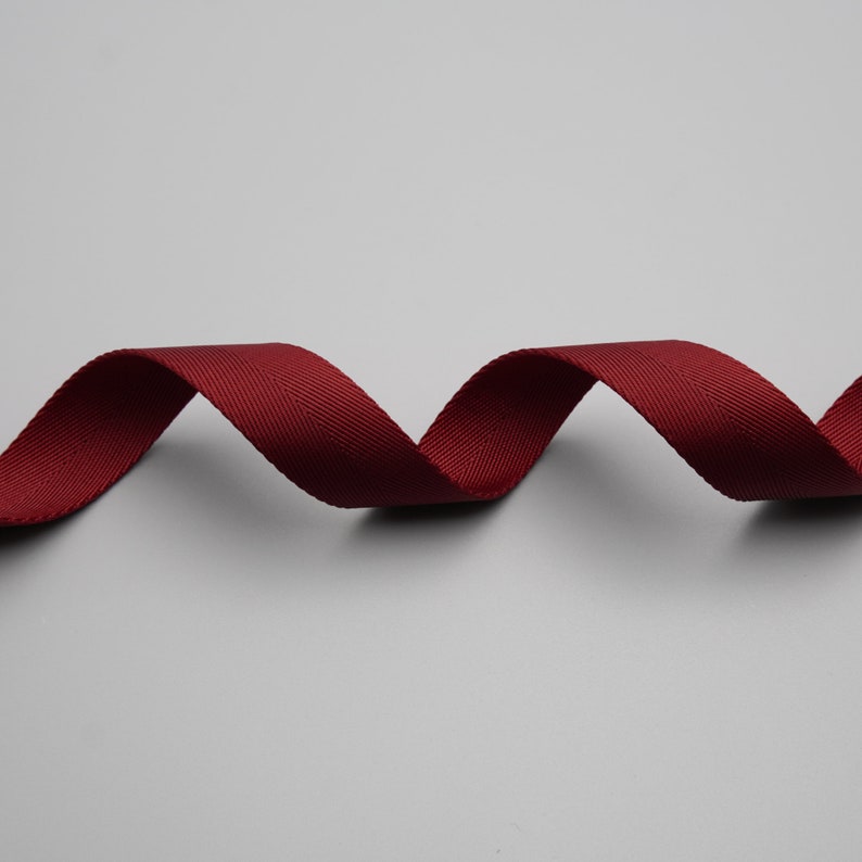 Peut inclure: Un ruban de couleur bordeaux, tiss&eacute; avec un motif diagonal subtil, ondule sur un fond gris. La texture du ruban est visible, sugg&eacute;rant un mat&eacute;riau durable. Le ruban est probablement utilis&eacute; pour l'artisanat, la couture ou &agrave; des fins d&eacute;coratives.