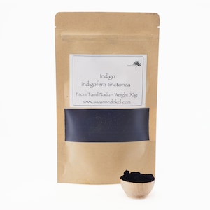 Può includere: Un sacchetto di carta marrone con un'etichetta che recita "Indigo indigofera tinctoria From Tamil Nadu - Weight 50gr www.suzannedekel.com". Il sacchetto contiene una polvere blu scuro. Una piccola ciotola di legno con una paletta di polvere è in primo piano.