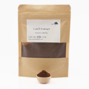 Peut inclure: Un sac en papier brun avec une &eacute;tiquette blanche qui indique "Cutck Extract Acacia Catechu poids : 50gr / 200gr www.suzannedekel.com". Le sac contient une poudre brune. Un petit bol en bois rempli de la poudre brune se trouve devant le sac.