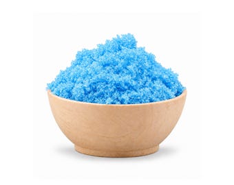 May include: A small, round, light-colored wooden bowl filled to the brim with a mound of vibrant, bright blue granulated crystals. The texture of the contents appears coarse and uniform, contrasting with the smooth, natural grain of the bowl.
