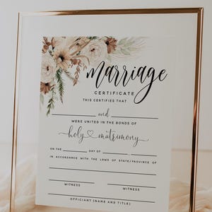 May include: A framed marriage certificate with elegant calligraphy. The certificate includes the words "Marriage Certificate" and "holy matrimony." Floral accents in neutral tones adorn the top left corner. The frame is gold.