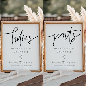 May include: Two gold framed signs with white backgrounds and black text. The sign on the left says "Ladies" and the sign on the right says "Gents". Both signs have the text "Please help yourself" and "Here is a basket to help you stay. Now hurry back out there before you miss the next song!"