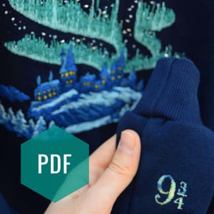 Könnte beinhalten: Marineblaues Sweatshirt mit gesticktem Schloss- und Nordlicht-Design. Das Ärmelbündchen zeigt die gelbe Zahl "9 3/4". Ein grünes Sechseck mit dem Wort "PDF" befindet sich in der unteren linken Ecke.