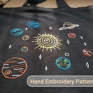 Könnte beinhalten: Eine schwarze Tragetasche mit einem handgestickten Design des Sonnensystems. Die Sonne befindet sich in der Mitte, umgeben von Planeten, Sternen und einem Ringplaneten. Der Text "Hand Embroidery Pattern" befindet sich am unteren Rand der Tasche.