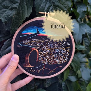 May include: Embroidered hoop art featuring a city night scene with a plane in the sky. The artwork uses black fabric and various colored threads to depict the city lights and a dark tunnel. The text "PDF PATTERN + TUTORIAL" is visible.