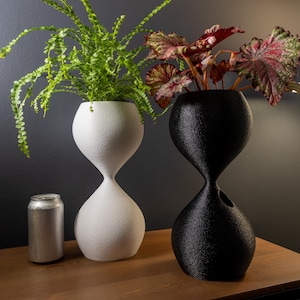 May include: Two hourglass-shaped planters, one white and one black, each holding greenery. The white planter has a textured surface, while the black one has a rougher texture. A silver can sits nearby. The planters are on a wooden surface.