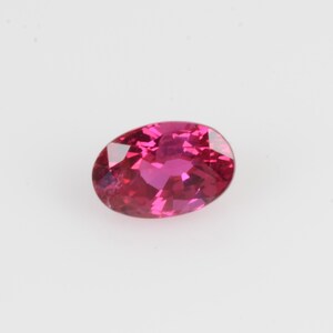 Puede incluir: Una gema facetada de forma ovalada en un vibrante tono magenta. La gema muestra claridad interna y una superficie pulida, reflejando la luz para crear un brillo brillante. La gema se encuentra sobre un fondo blanco liso.