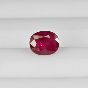 Puede incluir: Gema de rubí de corte ovalado con un tono rojo intenso. La gema tiene múltiples facetas que reflejan la luz, creando un efecto brillante. El rubí está sobre un fondo blanco.
