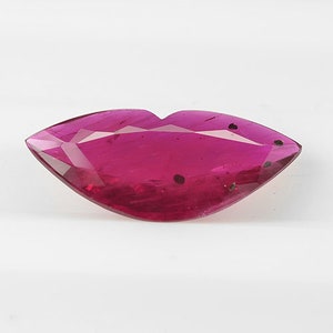 May include: A faceted, ruby-colored gemstone cut in the shape of lips. The gem has a high-gloss finish and is set against a white background. The gem has a few dark inclusions.