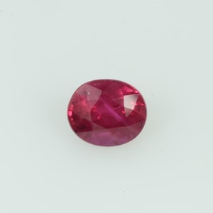May include: A faceted, oval ruby gemstone. The gem is a deep red colour with internal inclusions. The cut of the stone creates multiple reflective surfaces, enhancing its brilliance. The ruby is set against a plain white background.