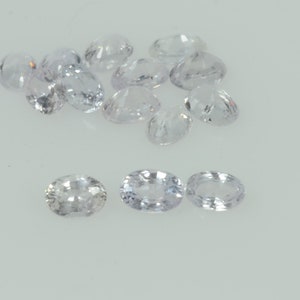 May include: A selection of oval-shaped, pale lavender gemstones. The gems are arranged on a white surface, with different sizes and facets. Light reflects off the cut surfaces, creating a sparkling effect. These gemstones are suitable for jewellery making.