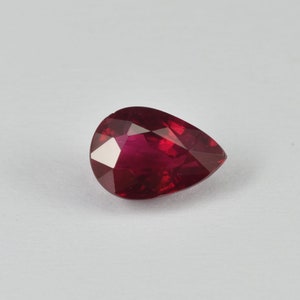 Pode incluir: Uma pedra preciosa de rubi facetada em forma de pêra. A gema é de uma cor vermelha profunda e rica com múltiplas facetas que refletem a luz. A pedra preciosa está sobre um fundo branco liso.
