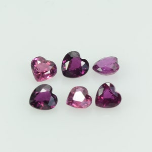 May include: Six heart-shaped gemstones in varying shades of pink and purple. The gemstones are faceted, with visible internal structures and light reflections. The arrangement is scattered against a white background, highlighting the gems' color and shape.