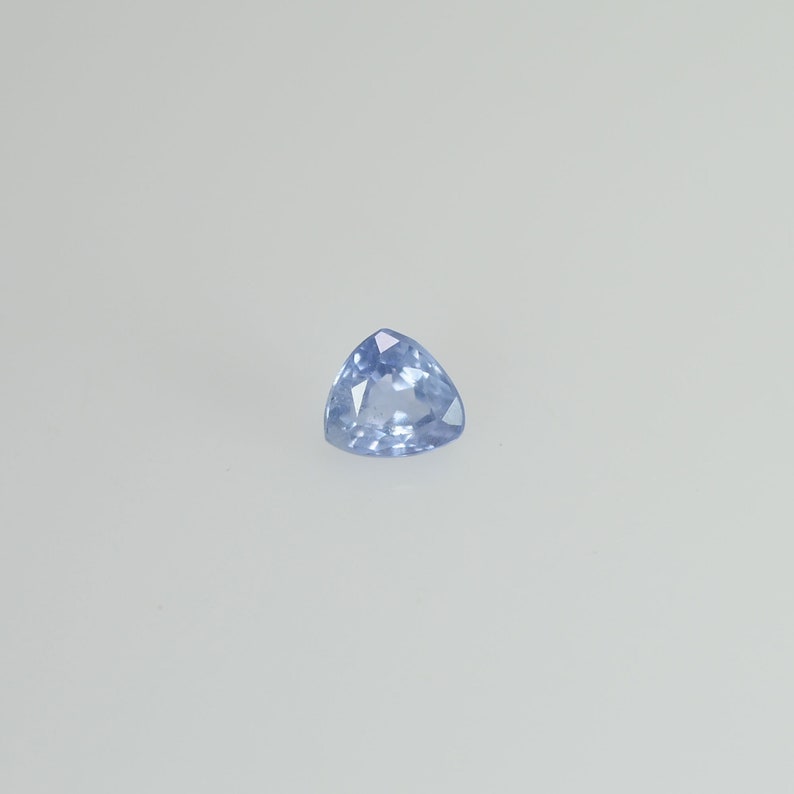 May include: A light blue, triangular gemstone with a faceted surface. The gem is set against a white background, showcasing its clarity and color. The gemstone's shape is a perfect equilateral triangle.