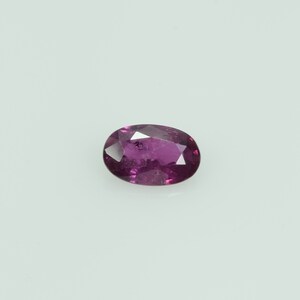 May include: An oval-cut, faceted gemstone in a deep magenta hue. The gem is set against a white background, showcasing its polished surfaces and internal clarity. The stone's shape and color suggest it could be a ruby or a similar precious stone.