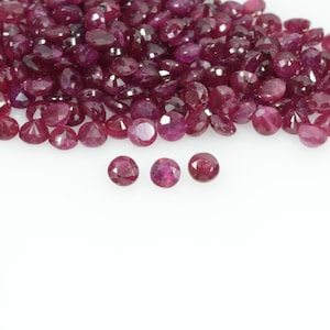 May include: A collection of faceted, round rubies in varying sizes and shades of red. The gemstones are scattered across a white surface, with three rubies prominently displayed in the foreground. These gems are ideal for jewelry making.