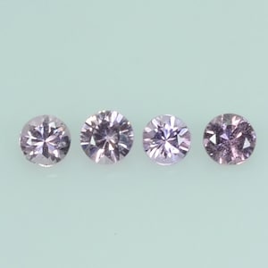 May include: Four round, light purple gemstones are arranged in a row against a light green background. Each gem has multiple facets that reflect light, creating a sparkling effect. The gems are of similar size and shape.