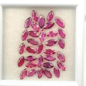 May include: A selection of marquise-cut gemstones in shades of pink and magenta, displayed on a white, textured background within a white frame. The gems are faceted, with a polished, reflective surface.