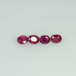 May include: Four oval-cut, deep red rubies are displayed against a white background. The gemstones are arranged in a horizontal line, showcasing their polished surfaces and internal brilliance. The rubies vary slightly in size and shade.
