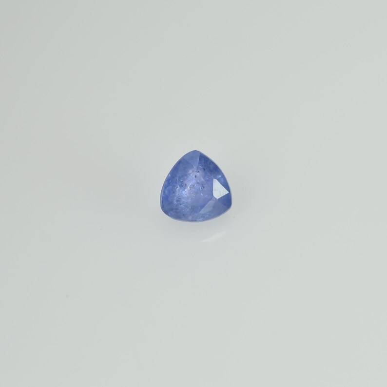 May include: A faceted, triangular blue gemstone. The gem has a light blue hue with a polished surface and sharp edges. The gemstone is set against a white background, highlighting its color and shape.
