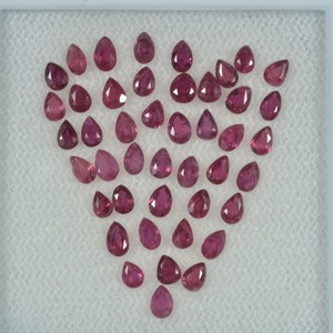 May include: A collection of pear-shaped, deep red rubies arranged in a heart shape. The gemstones are displayed on a white, textured surface within a white frame. Each ruby has a polished, faceted surface.