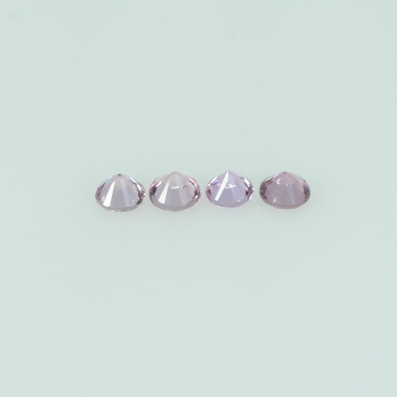May include: Four round, faceted gemstones in a row. The gems are a light purple color and appear to be cut with many facets. The background is a solid, light-colored surface.