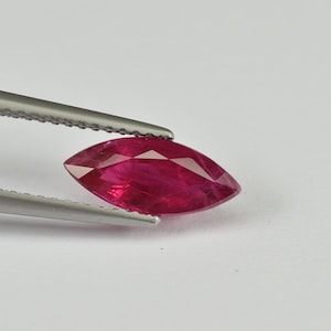 May include: A marquise-cut ruby gemstone, held by silver tweezers. The ruby is a deep red colour, with visible internal inclusions. The gemstone is displayed against a plain white background, highlighting its shape and colour.