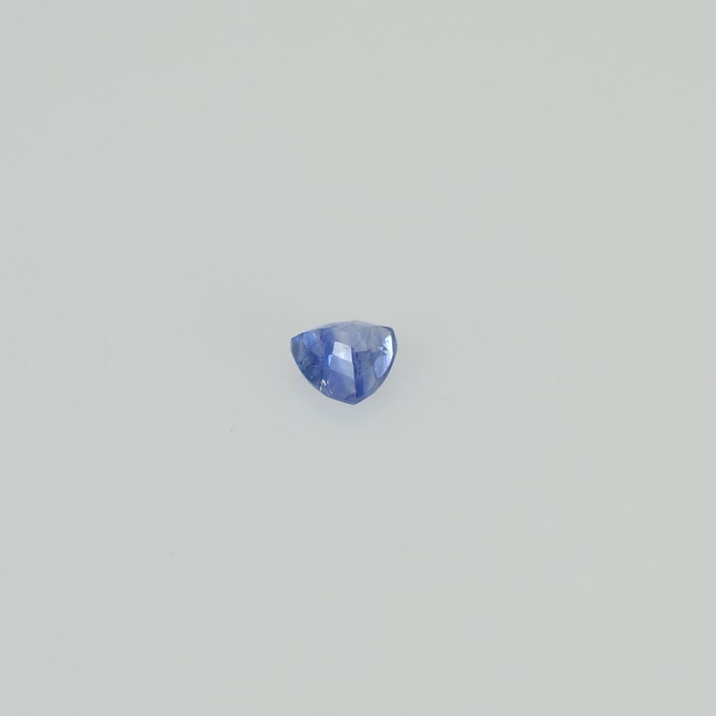 May include: A faceted, triangular blue gemstone. The gem has a light blue hue with a slightly darker blue at the edges. The stone is set against a plain white background.