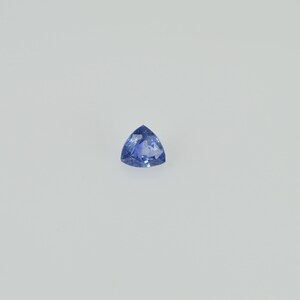 May include: A triangular, faceted, blue gemstone. The gem has a brilliant cut, with a deep, saturated color. The stone is set against a plain white background, highlighting its shape and color.