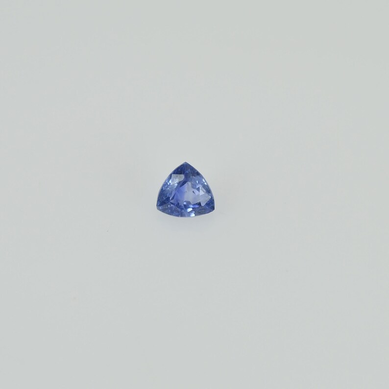 May include: A triangular, faceted, blue gemstone. The gem has a brilliant cut, with a deep, saturated color. The stone is set against a plain white background, highlighting its shape and color.