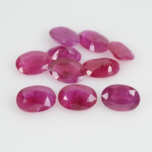 May include: A collection of oval-cut rubies, showcasing a deep red hue. The gemstones are arranged on a white surface, with facets catching the light. The rubies vary slightly in size and shade, highlighting their natural beauty.