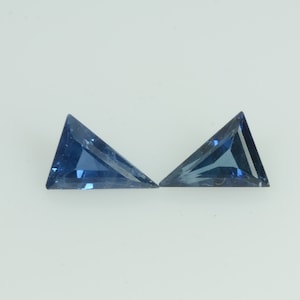 Puede incluir: Dos gemas triangulares facetadas de color azul intenso. Las gemas se muestran sobre un fondo blanco, resaltando sus ángulos agudos y superficies reflectantes. Las gemas probablemente están talladas para joyería.