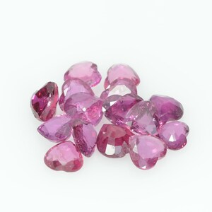 Puede incluir: Un conjunto de piedras preciosas en forma de corazón en tonos rosa y magenta. Las piedras facetadas están dispersas sobre una superficie blanca, con la luz reflejándose en sus superficies pulidas. Las piedras preciosas son de diferentes tamaños.