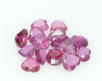 May include: A cluster of heart-shaped gemstones in shades of pink and magenta. The faceted stones are scattered on a white surface, with light reflecting off their polished surfaces. The gemstones are of different sizes.