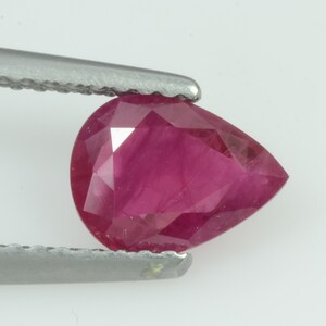 May include: A faceted, pear-shaped ruby gemstone held by tweezers. The ruby is a deep, rich red color, with multiple facets catching the light. The gemstone is a teardrop shape, and the tweezers are silver.