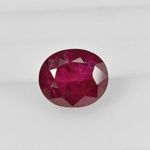 Puede incluir: Una gema de rubí tallada en forma ovalada y facetada. La gema es de un color rojo intenso y rico, con múltiples facetas que reflejan la luz. La piedra preciosa se encuentra sobre un fondo blanco.