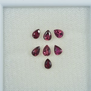 May include: Seven pear-shaped, deep red gemstones are displayed on a white, textured surface within a white frame. The gems are arranged in a scattered pattern. Each stone has a polished surface, reflecting light and showcasing their colour.