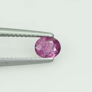 May include: A faceted, oval-shaped pink gemstone is held by a pair of silver tweezers against a white background. The gemstone has a slightly irregular shape and internal inclusions, with a polished surface that reflects light.