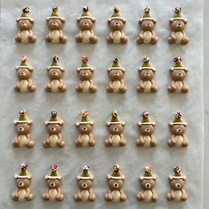 May include: Twenty-four small, brown teddy bear figurines are arranged in a grid. Each bear wears a green party hat adorned with colourful sprinkles. The bears are on a light-coloured surface.