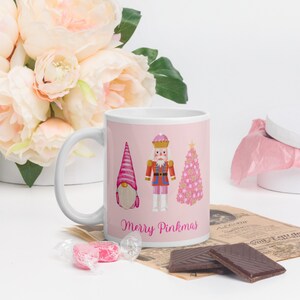 Op de afbeelding: Witte keramische mok met een roze achtergrond waarop een roze kabouter, een roze kerstboom en een roze notenkraker te zien zijn. De tekst "Merry Pinkmas" staat in roze op de mok.