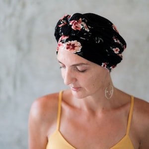 Può includere: Una donna con un turbante a stampa floreale sulla testa. Il turbante è nero con fiori rosa e bianchi. Indossa una canotta gialla e un orecchino d'argento.