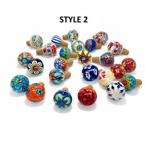Può includere: Una collezione di tappi per bottiglie decorativi in ceramica con vari disegni floreali e geometrici colorati. I tappi hanno basi in sughero e sono dipinti in tonalità di blu, rosso, giallo e verde. Il testo "STYLE 2" è in alto.