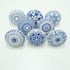 May include: Eight white ceramic cabinet knobs with blue decorative patterns. The knobs display diverse geometric and floral designs, suitable for furniture or cabinetry. Each round knob has a metal screw for easy fitting. The knobs are approximately 4 cm in diameter.