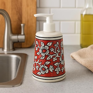 May include: A red ceramic soap dispenser with a white pump. The body features a white floral pattern. Black stripes border the top and bottom. The dispenser is on a grey countertop near a stainless steel sink.