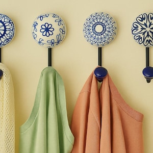 May include: Four decorative wall hooks with blue and white patterned ceramic knobs. The hooks are black with a blue knob at the bottom. Various coloured items are hanging from the hooks, including a yellow mesh scarf and green and peach shirts.