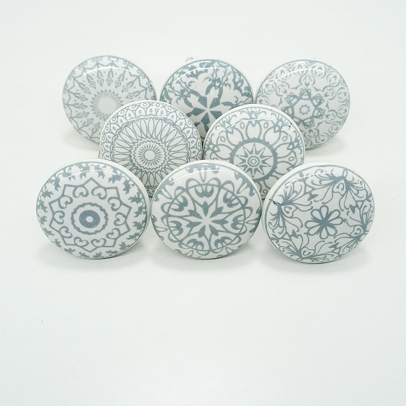 May include: Eight white ceramic cabinet knobs with detailed grey floral and geometric designs. The knobs are round and have a glossy finish, ideal for adding a decorative touch to furniture or cabinets. Each knob has a diameter of approximately 4 cm.