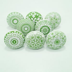 Könnte beinhalten: Acht weiße Keramik-Schrankknöpfe mit grünen geometrischen Mustern. Die Knöpfe weisen kreisförmige Designs auf, darunter Blumen, Mandalas und abstrakte Formen. Geeignet für Möbel oder Dekoration.