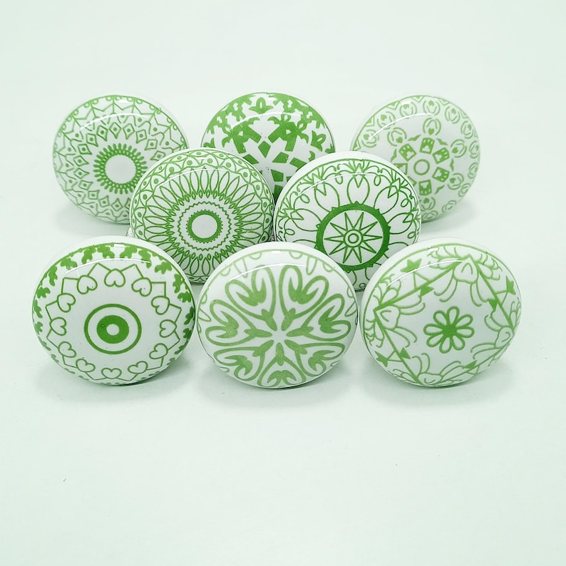 May include: Eight white ceramic cabinet knobs with green geometric patterns. The knobs feature circular designs, including floral, mandala, and abstract shapes. Suitable for furniture or decor, each knob has a unique design.