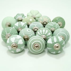 以下が含まれることがあります： 白のフローラルと幾何学模様が施された、セージグリーンのセラミック製キャビネットノブのコレクション。各ノブはシルバー色のベースです。ノブは形状とデザインが異なり、さまざまな質感とスタイルを提供します。
